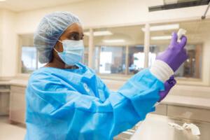 全球最大的博彩平台. Dunn School of 药店 student in blue scrubs, gloves, cap and mask, holding a vial.